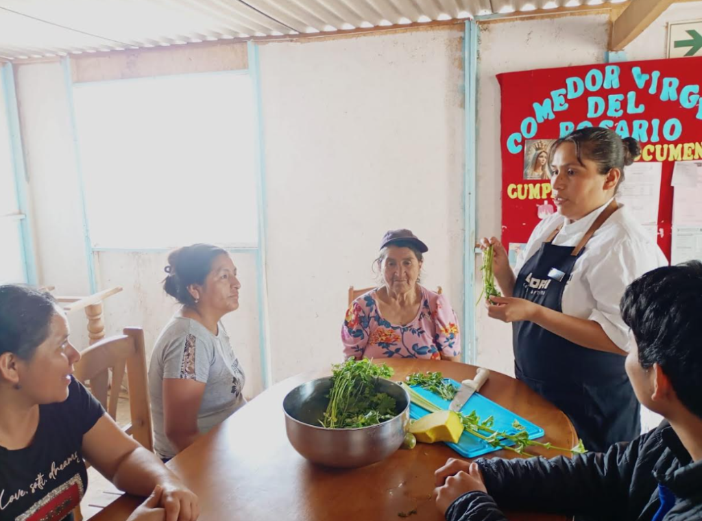 Mujeres participan en una clase de cocina, observando a la anfitriona que sostiene hierbas frescas. En una mesa, hay un bol con verduras y utensilios de cocina. Al fondo, se ve un cartel del comedor comunitario.