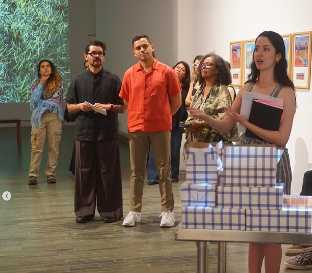 Una mujer habla frente a un grupo de personas en una galería de arte, con una presentación proyector proyectada de fondo y cajas de regalo decorativas en una mesa.