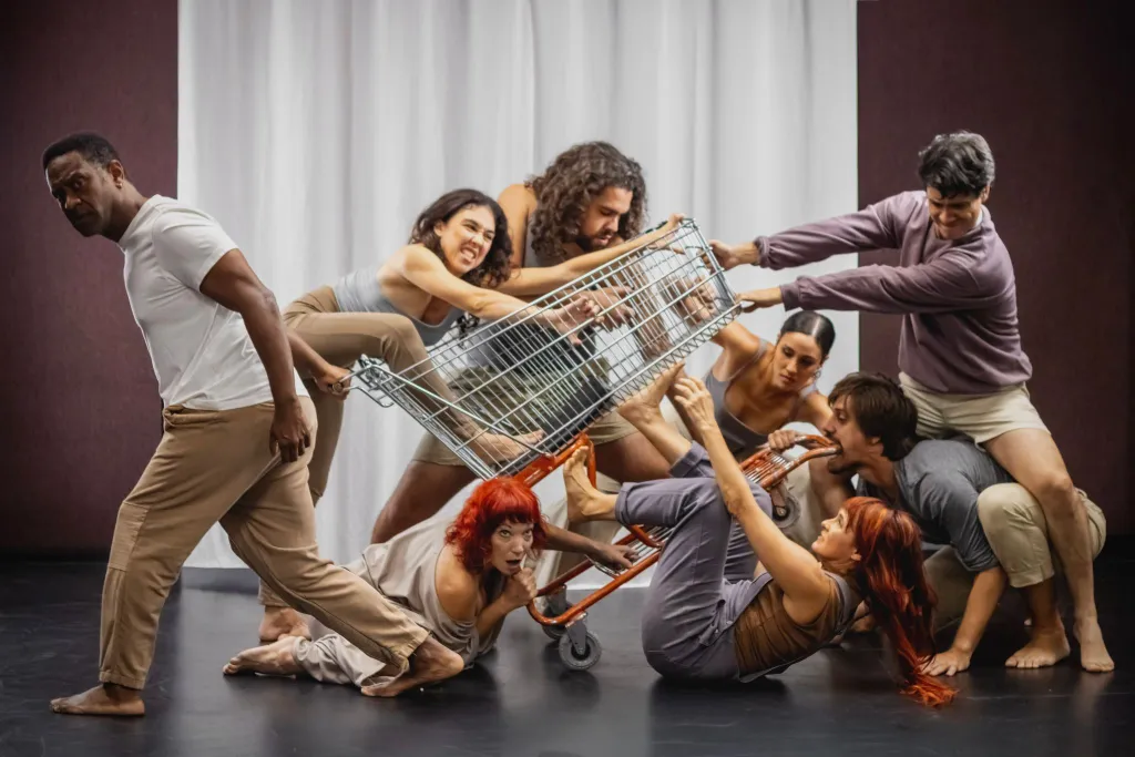 Un grupo de ocho personas interactuando con un carrito de supermercado en una rutina de baile, con expresiones faciales diversas que reflejan energía y dinamismo.